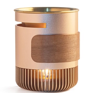 Può includere: Un portacandele cilindrico con finitura metallica color oro rosa, una fascia decorativa in legno liscio avvolta attorno al centro e una base composta da listelli verticali che rivelano una piccola candela accesa all'interno.