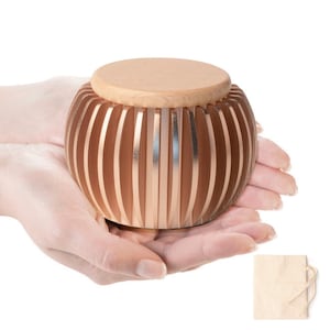 Puede incluir: Un recipiente decorativo de madera redondo con tapa marrón claro. El recipiente tiene listones verticales de color cobre. El recipiente se sostiene con las manos abiertas. Una pequeña bolsa de papel beige está en la esquina inferior derecha.