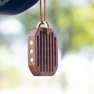 Op de afbeelding: Een houten auto luchtverfrisser met een goudkleurige metalen sluiting en een bruin koord. De rechthoekige verfrisser heeft verticale groeven en drie witte cirkelvormige accenten. De achtergrond is wazig.