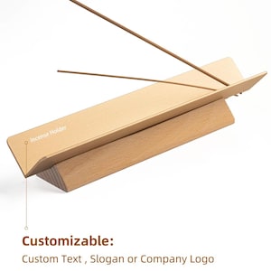 May include: Wooden incense holder with a slanted top and a triangular base. The holder has the text "Incense Holder" printed on the top. Two incense sticks are placed on the holder.