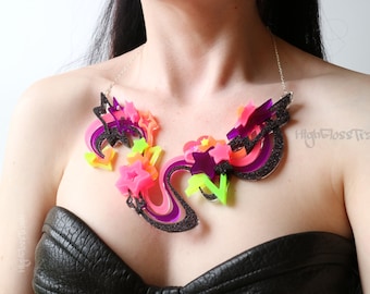 May include: A vibrant statement necklace featuring neon pink, orange, and green star and swirl designs. The necklace has a black glitter border and a silver chain. The text "HighGlossTrash" is visible.