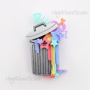 Pode incluir: Uma lata de lixo prateada com glitter com um arco-íris de estrelas, corações e ossos que se derramam pela parte superior. O texto "High Gloss Trash" é visível no fundo.
