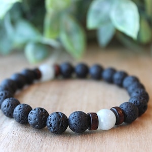 Könnte beinhalten: Ein Armband aus schwarzen Lavasteinperlen mit weißen und braunen Akzenten. Das Armband besteht aus runden, porösen schwarzen Perlen, die mit weißen und Holzperlen durchsetzt sind. Das Armband wird auf einer Holzoberfläche präsentiert.