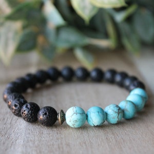 May include: A beaded bracelet featuring a combination of black lava rock beads and turquoise beads. The bracelet has a mix of textures and colors, with the black beads on one side and the turquoise beads on the other. The bracelet is on a wooden surface.