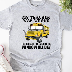 Puede incluir: Camiseta gris claro con un dibujo de autobús escolar amarillo. El texto de la camiseta dice: "MY TEACHER WAS WRONG I DO GET PAID TO STARE OUT THE WINDOW ALL DAY."