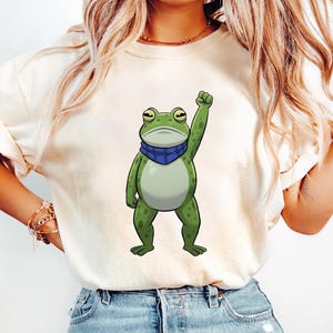 以下が含まれることがあります： クリーム色のTシャツ。漫画のキャラクターであるカエルが拳を突き上げている。カエルは緑色で、青いスカーフを着用。ゆったりとしたフィット感で、半袖。カエルのグラフィックはTシャツの中央に配置されている。
