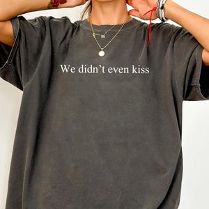 May include: Dark grey oversized t-shirt with the text "We didn't even kiss" in white. The shirt has short sleeves and a relaxed fit. The shirt is made of a soft, comfortable material.