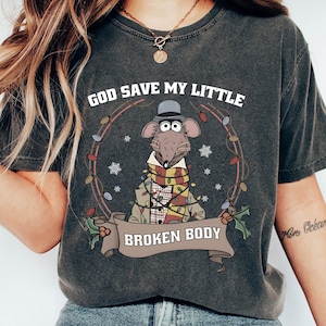 May include: Dark gray t-shirt featuring a cartoon rat wearing a hat, scarf, and Christmas lights. The text "GOD SAVE MY LITTLE" is above the rat, and "BROKEN BODY" is below. The design is surrounded by a wreath.