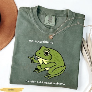 May include: Moss green t-shirt with a cartoon frog graphic. The frog is green with a yellow belly and is pointing. Text on the shirt reads "me: no problemo!" and "narrator: but it was all problemo". A gold necklace is draped on the shirt.
