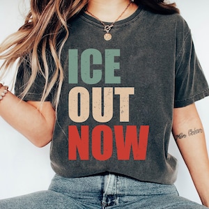May include: Dark gray t-shirt with the words "ICE OUT NOW" in teal, beige, and red block letters. The shirt is a casual style, suitable for everyday wear.