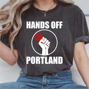 May include: Dark gray t-shirt with the text "HANDS OFF PORTLAND" in white. The design features a white fist holding a red rose inside a white circle. The shirt is a casual wear item.