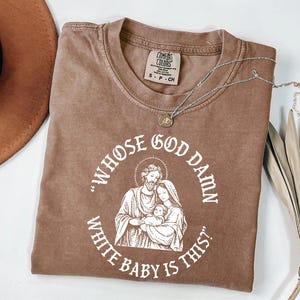 May include: Brown Comfort Colors t-shirt featuring the text "WHOSE GOD DAMN WHITE BABY IS THIS?" encircling a white illustration of a family. A silver necklace with a pendant rests on the shirt.