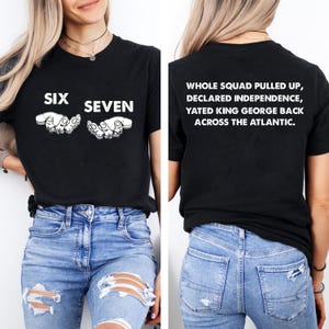 May include: Black t-shirt with white text and graphics. The front of the shirt says "SIX SEVEN" with hand illustrations. The back of the shirt says "WHOLE SQUAD PULLED UP, DECLARED INDEPENDENCE, YATED KING GEORGE BACK ACROSS THE ATLANTIC."