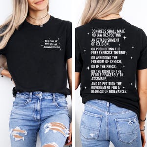 May include: Black t-shirt with white text on the front and back. The front says "Big fan of the First Amendment." The back lists the First Amendment's clauses. The shirt is paired with blue jeans.