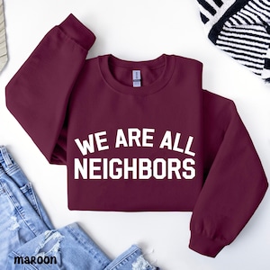 Op de afbeelding: Bordeauxkleurige crewneck-trui met de witte tekst "WE ARE ALL NEIGHBORS". De trui wordt gepresenteerd op een witte ondergrond, met een spijkerbroek en een zwart-wit patroon sjaal op de achtergrond.
