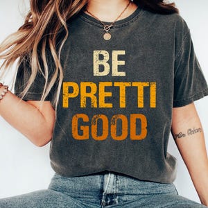 May include: Dark gray t-shirt with the words "BE PRETTI GOOD" in distressed yellow and orange lettering. The shirt has a vintage, worn look.