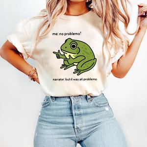 May include: Cream-colored t-shirt featuring a cartoon frog with the text "me: no problemo!" and "narrator: but it was all problemo". The frog is green with black outlines and is pointing with one hand. The shirt is paired with blue jeans.