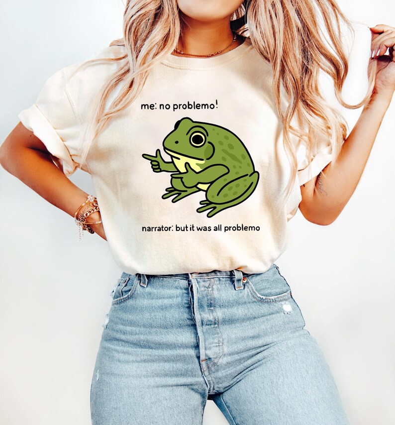 May include: Cream-colored t-shirt featuring a cartoon frog with the text "me: no problemo!" and "narrator: but it was all problemo". The frog is green with black outlines and is pointing with one hand. The shirt is paired with blue jeans.