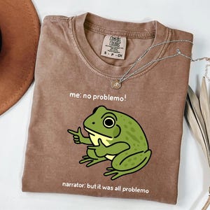 May include: A brown t-shirt with a green frog graphic pointing its finger. The text on the shirt reads "me: no problemo!" and "narrator: but it was all problemo". A silver necklace with a pendant is on the shirt.