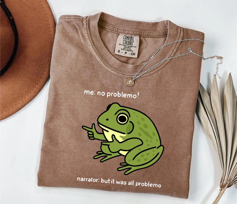May include: A brown t-shirt with a green frog graphic pointing its finger. The text on the shirt reads "me: no problemo!" and "narrator: but it was all problemo". A silver necklace with a pendant is on the shirt.