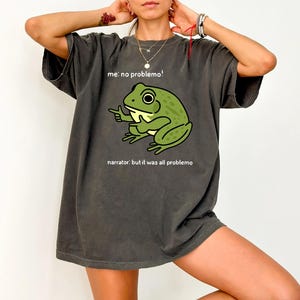 May include: Dark gray t-shirt featuring a cartoon frog with the text "me: no problemo!" above and "narrator: but it was all problemo" below. The frog is green and pointing with one hand.