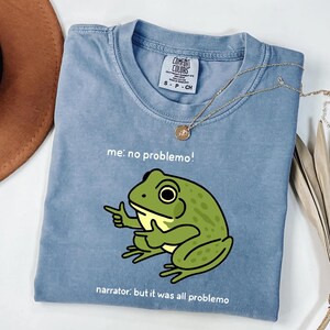 May include: A light blue Comfort Colors t-shirt with a green frog graphic pointing its finger. The text "me: no problemo!" and "narrator: but it was all problemo" are printed on the shirt. A gold necklace is draped across the shirt.