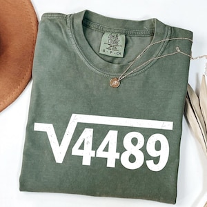Può includere: Una maglietta verde oliva con la scritta "√4489" in bianco. La maglietta ha un girocollo e maniche corte. Una collana d'oro con un ciondolo è drappeggiata sulla maglietta.