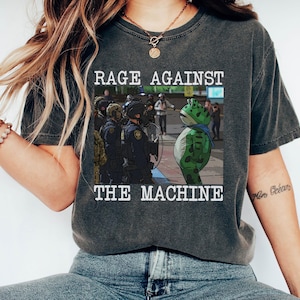 Peut inclure: T-shirt gris foncé avec l'inscription "RAGE AGAINST THE MACHINE" au-dessus et en dessous d'une image de grenouille de dessin animé face à des policiers. La grenouille est verte avec des taches noires et une écharpe bleue.