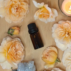 Könnte beinhalten: Eine dunkelbraune Glasflasche mit schwarzem Verschluss, umgeben von pfirsichfarbenen Rosen und einem Kristallhaufen. Die Flasche steht auf einer hellbraunen Holzoberfläche.