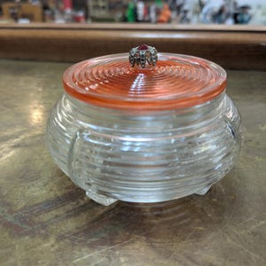 May include: A clear glass jar with horizontal ribbed detailing and three small feet. The lid is a translucent orange color with concentric circular ridges and a central silver-toned metal finial featuring a small, dark red stone accent.