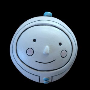 May include: A white, round ceramic face with black outlines and a smiling expression. It has a small nose, two black dots for eyes, and pink circles for cheeks. A small turquoise bead is on top and at the bottom.