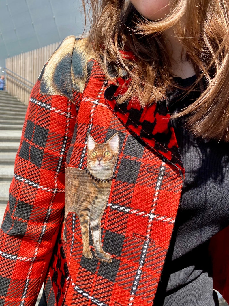 May include: A red and black plaid jacket with a cat applique. The cat is brown and black with a black collar.