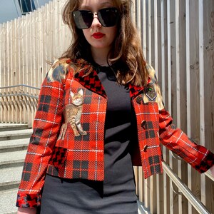 May include: A red and black plaid cropped jacket with a cat print on the front. The jacket has a velvet collar and a gold button closure.