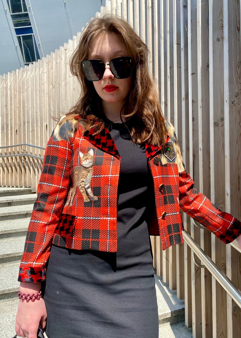 May include: A red and black plaid cropped jacket with a cat print on the front. The jacket has a velvet collar and a gold button closure.