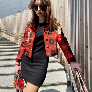 May include: A woman wearing a red and black plaid jacket with a black dress and a small red handbag. She is wearing sunglasses and has long brown hair.