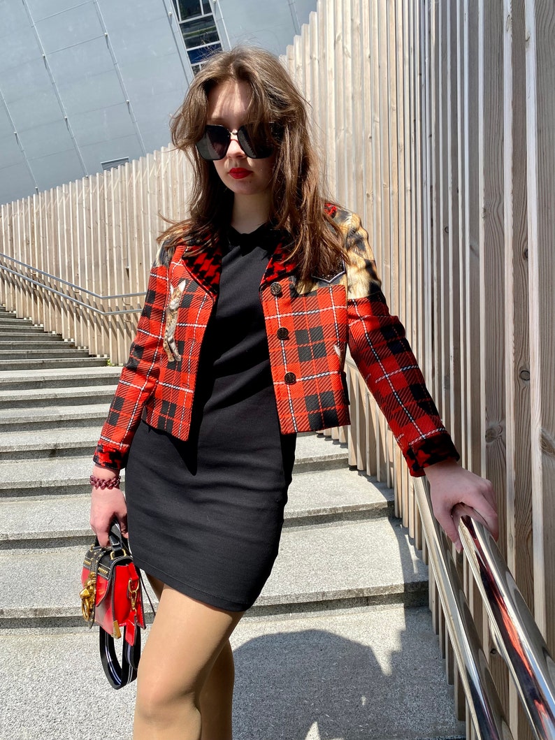 May include: A woman wearing a red and black plaid jacket with a black dress and a small red handbag. She is wearing sunglasses and has long brown hair.
