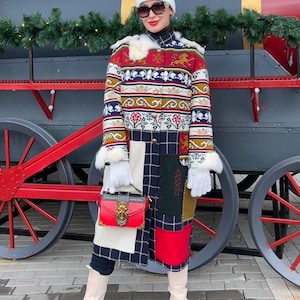 May include: A woman wearing a white beanie, sunglasses, and a long, colorful patchwork coat with fur trim. She is carrying a red handbag with a lion head clasp and is standing in front of a red and gray train car.