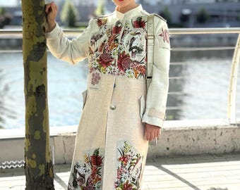 May include: A long, cream-colored coat with floral embroidery in red, white, and green. The coat features a high collar, long sleeves, and a single-button closure. The embroidery is concentrated on the upper chest and lower panels. The person is wearing sunglasses.