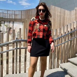 May include: A woman wearing a red and black plaid cropped jacket with a cat print, a black mini skirt, and black heeled shoes. She is standing on a set of stairs with a wooden railing.
