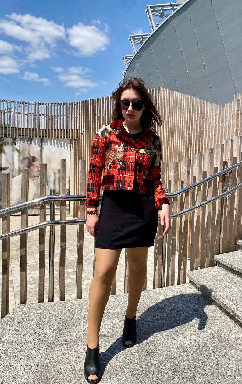 May include: A woman wearing a red and black plaid cropped jacket with a cat print, a black mini skirt, and black heeled shoes. She is standing on a set of stairs with a wooden railing.