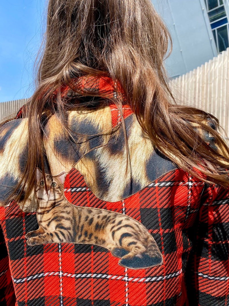 May include: A red and black plaid shirt with a brown and black cat applique. The cat is lying down on the shirt and has a white stripe on its forehead.