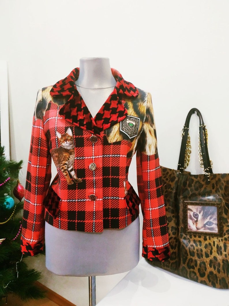 May include: A red and black plaid blazer with a black and white houndstooth collar. The blazer has a cat print on the front and a gold button closure. A leopard print tote bag with a gold chain strap is in the background.