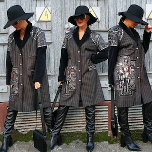 May include: A long, black and gray striped coat with a black velvet collar and black sleeves. The coat has a decorative embroidered panel on the front with a medieval scene. The model is wearing a black hat and black boots.