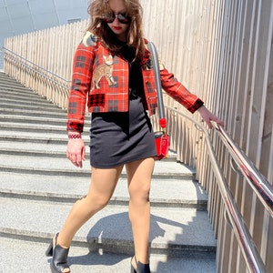 May include: A woman wearing a red and black plaid jacket with a cat print, a black mini dress, black heeled shoes, and sunglasses. She is walking down a set of stairs.