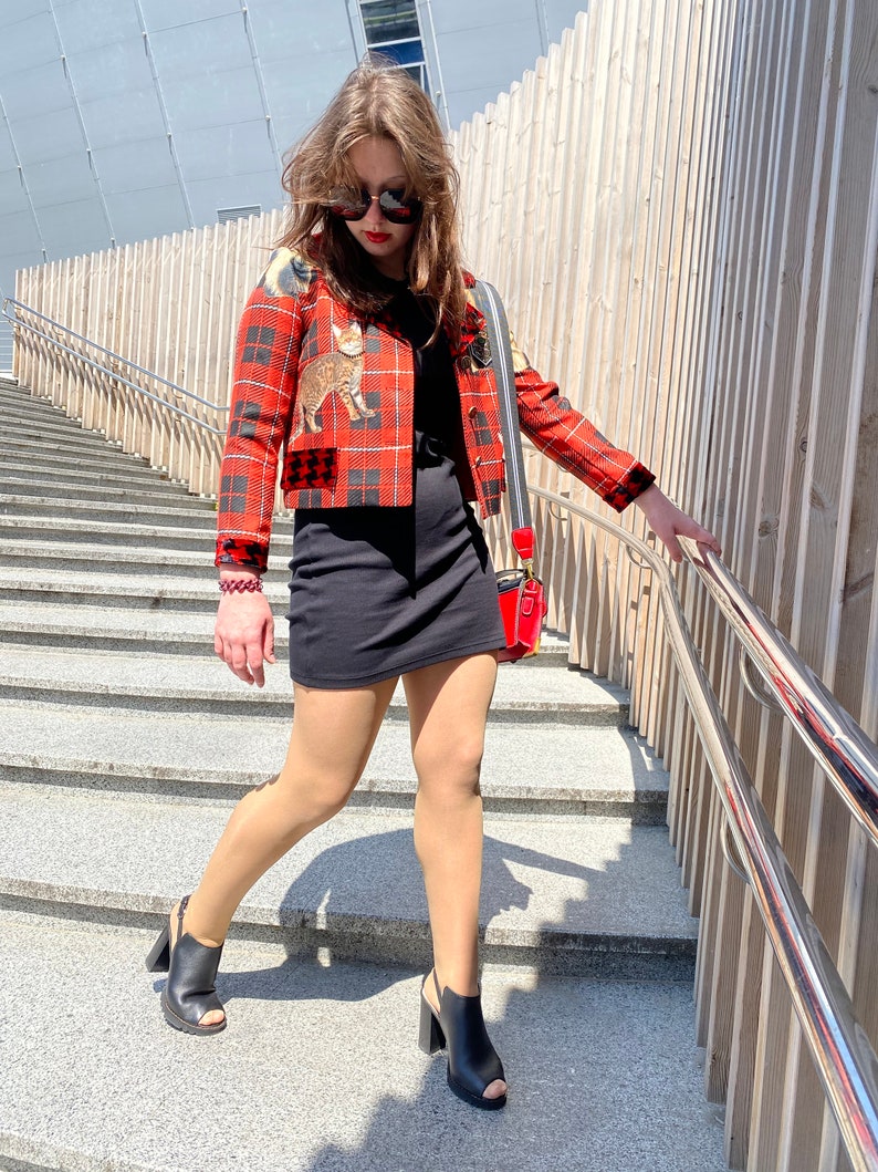 May include: A woman wearing a red and black plaid jacket with a cat print, a black mini dress, black heeled shoes, and sunglasses. She is walking down a set of stairs.
