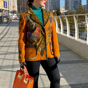 May include: A woman wearing a mustard yellow blazer with a colourful embroidered design, black trousers, and sunglasses. She is carrying an orange handbag.
