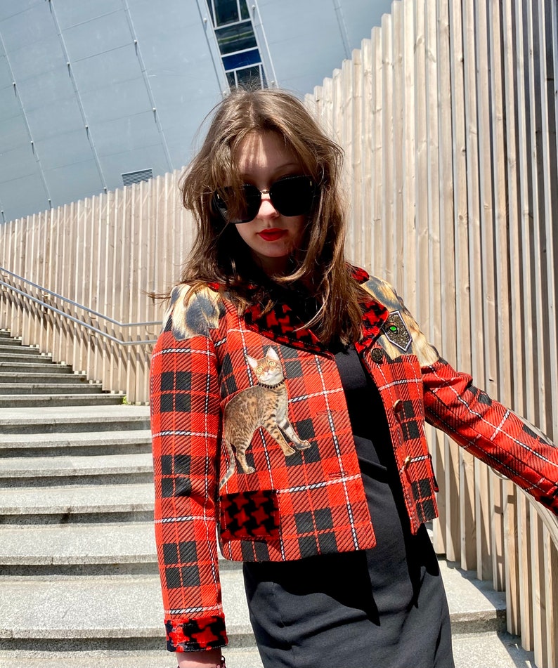 May include: A red and black plaid cropped jacket with a cat print patch and a black and white houndstooth pocket. The jacket has a black collar and a black zipper.