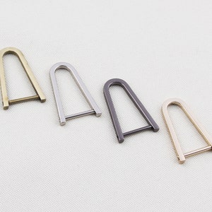 Op de afbeelding: Vier metalen U-vormige hardwarestukken in goud, zilver, gunmetal en messing tinten. Elk stuk heeft een rechthoekige basis en een afgeronde bovenkant, geschikt voor het maken van tassen of andere knutselprojecten.