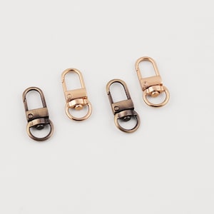 May include: Four metal swivel clasps in bronze and rose gold. Each clasp has a D-ring and a spring-loaded gate. The clasps are arranged on a white surface, suitable for crafting or jewellery making.