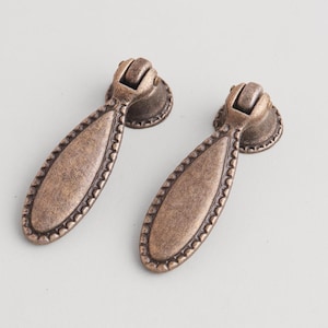May include: Two antique bronze-colored drawer pulls. Each pull has a teardrop-shaped handle with a beaded edge and a decorative backplate. The pulls are designed for furniture and cabinets.